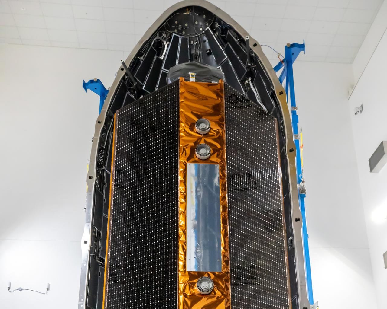A satellite undergoing payload integration in a launch processing facility — solar arrays, thermal blanket, and electronics enclosure visible.